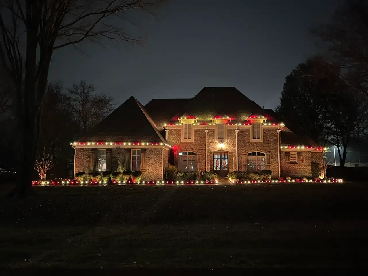 Brick two-story home with red and warm white C9 lights along the roofline and flowerbeds at dusk