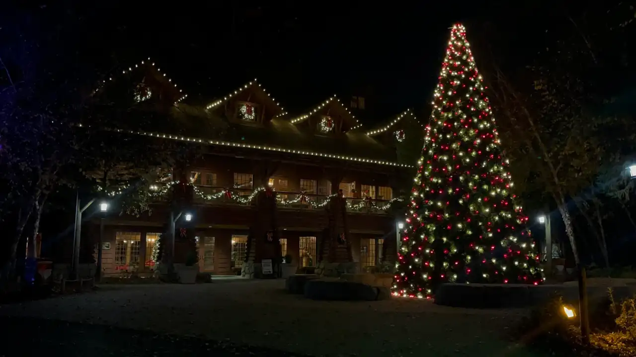 Lodge-style building outlined in warm white roofline lights and lit garland beside a large red and white Christmas tree