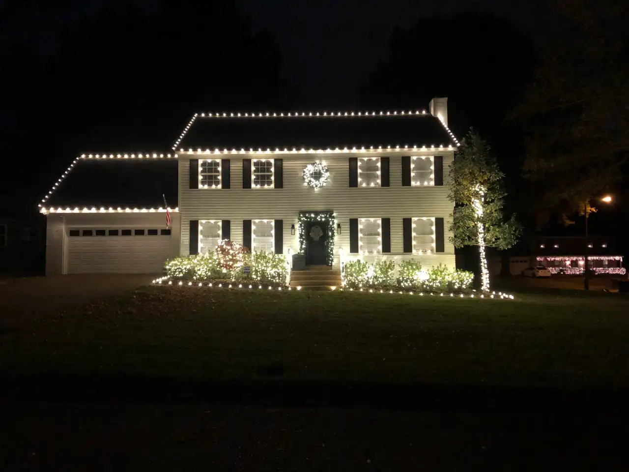 Colonial home with warm white roofline and window outlines, a lit wreath and garland around the door
