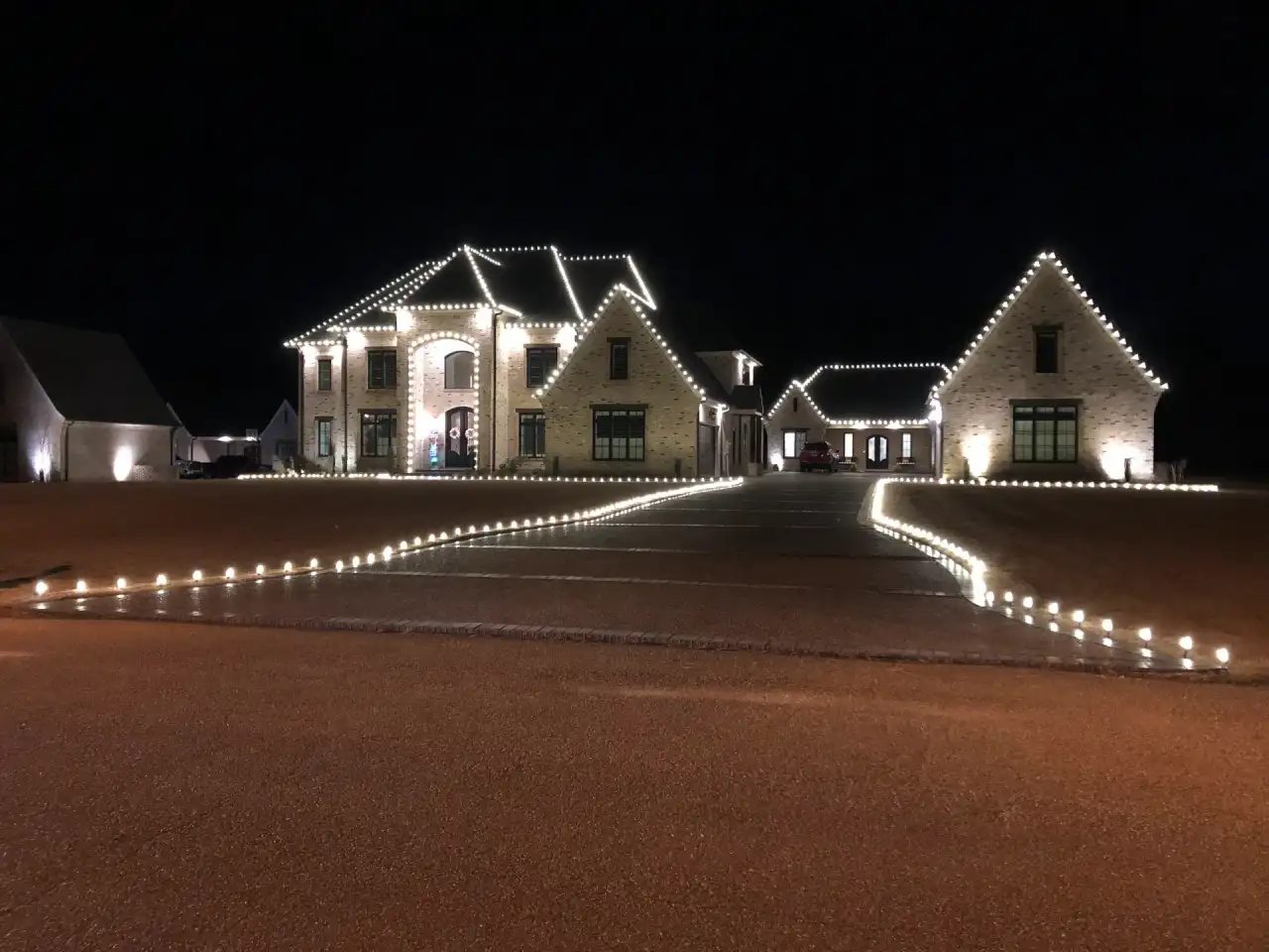 Large white-brick home with warm white roofline lights and lit driveway markers