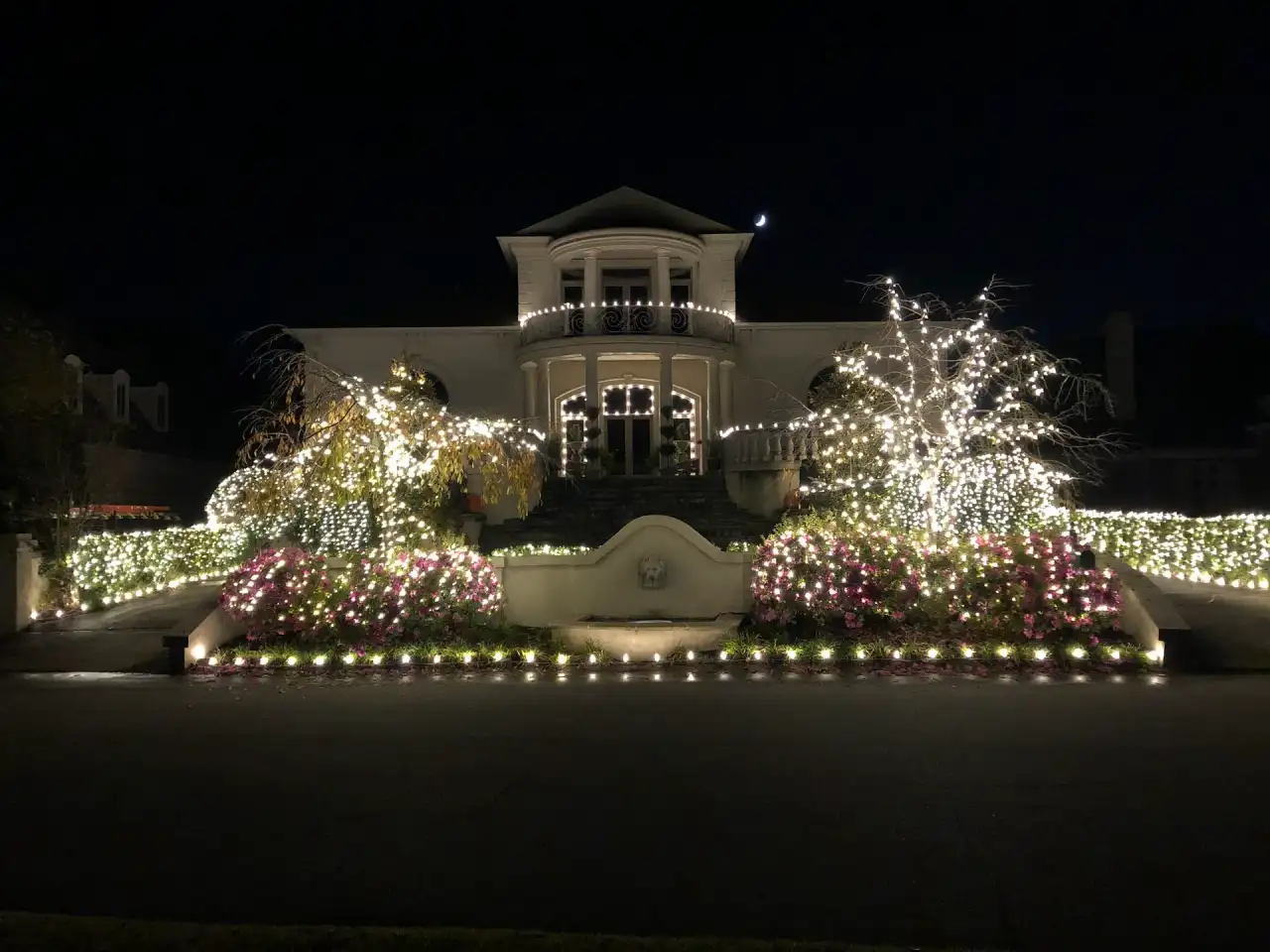Two-story home with trees, shrubs and flowerbeds wrapped in warm white lights at night