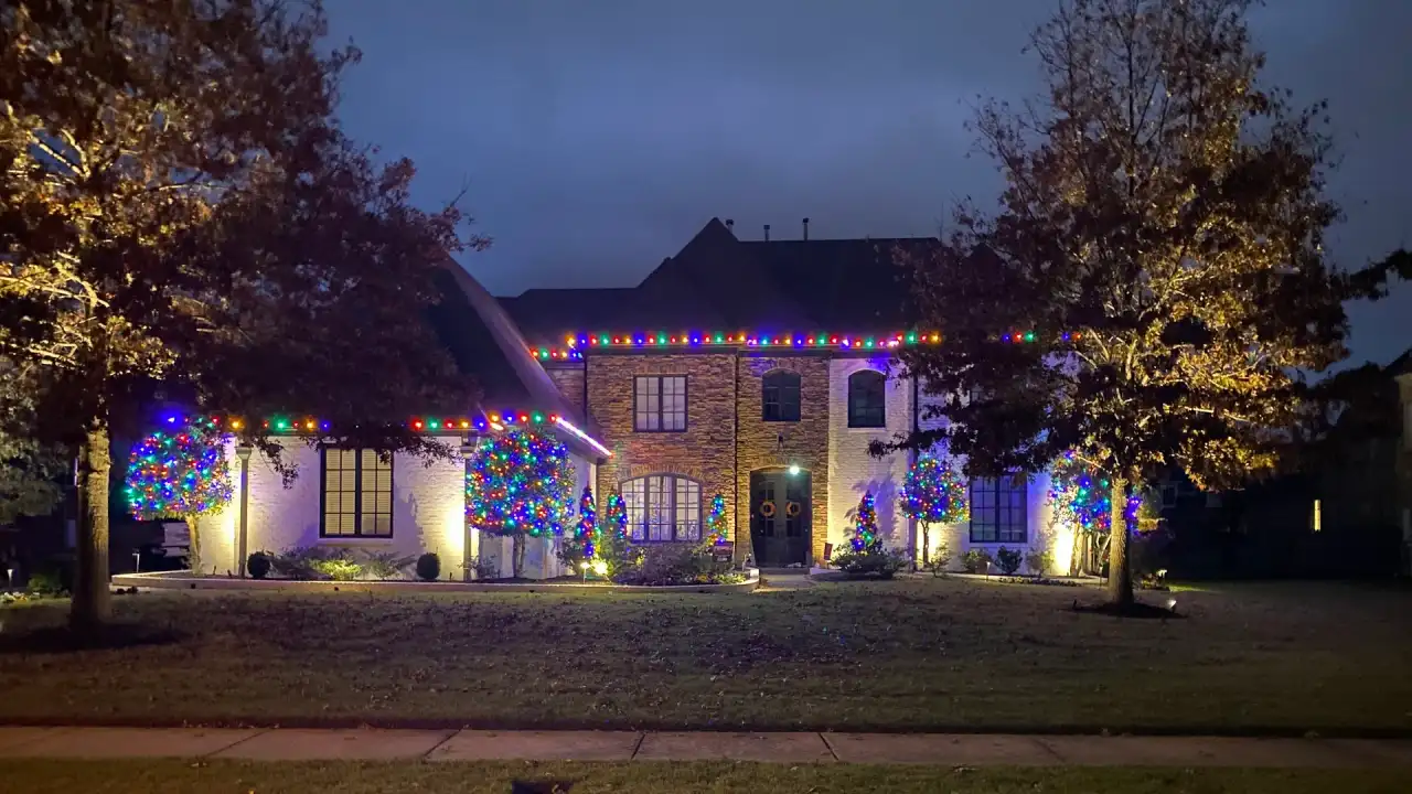 Brick home with multicolor lights on the roofline, shrubs and trees at dusk